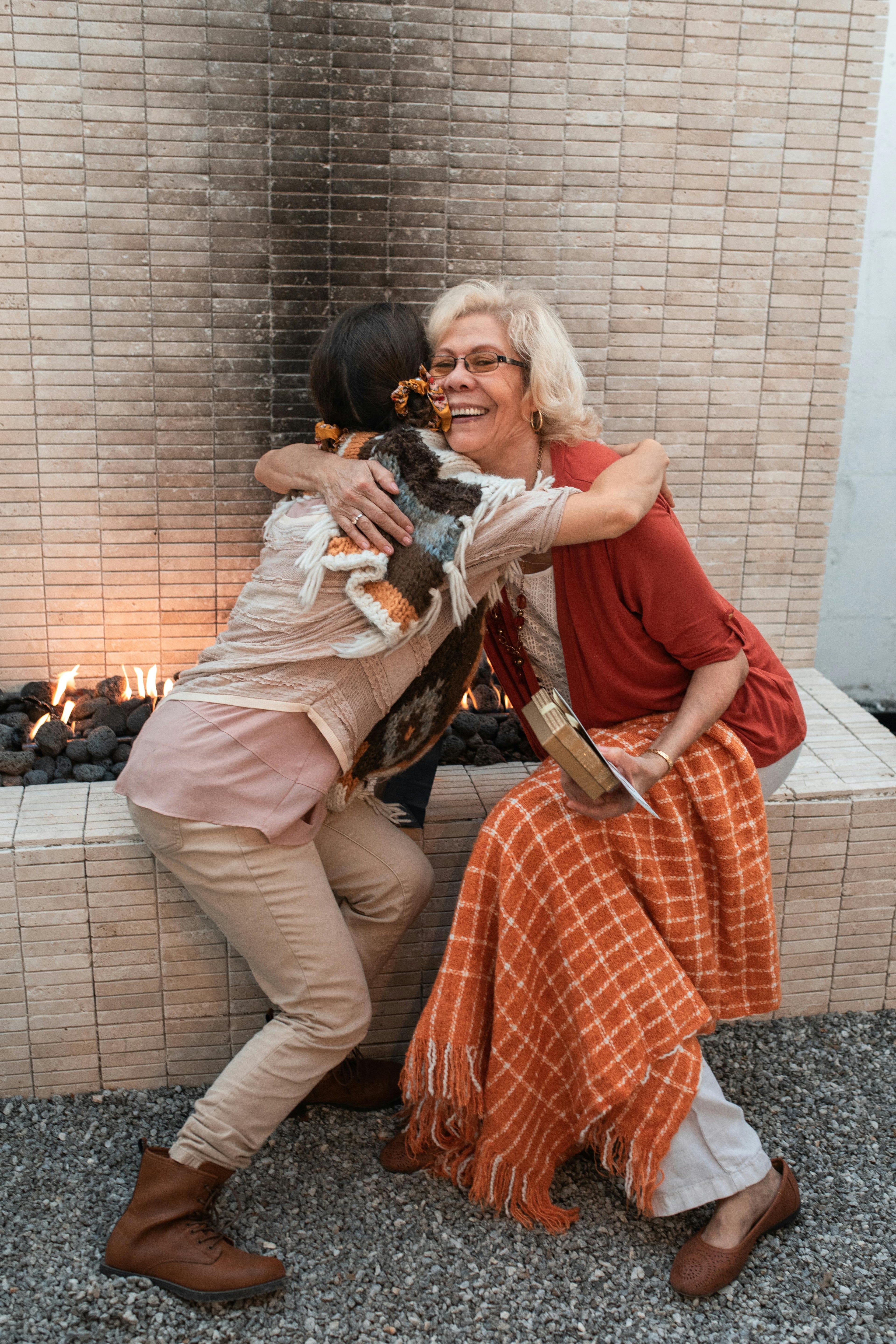 Two women hugging in front of a fireplace with a warm, cozy atmosphere.